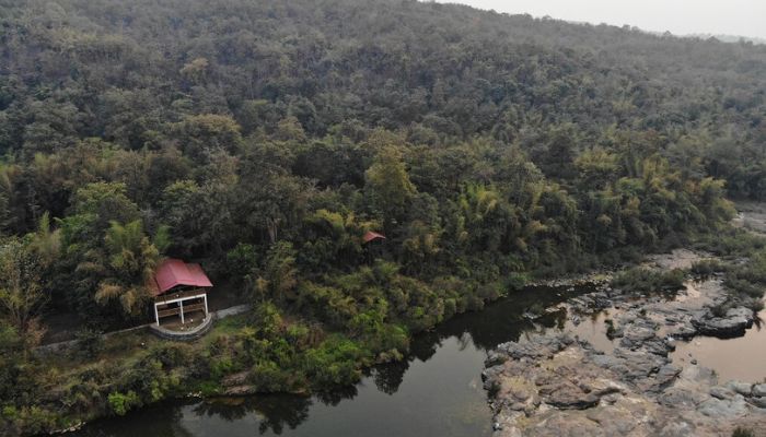 Dense forest surrounds the Mahal Eco Campsite with a small cabin near a rocky riverside